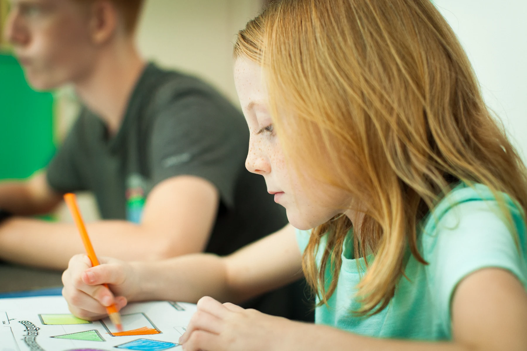 A child coloring a worksheet at a classroom table, with another student beside them.